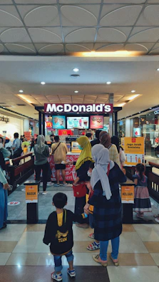 A busy McDonald's outlet in a shopping mall with several people forming a queue at the counter. The entrance and exit are marked with signs in Indonesian. Various digital menu boards displaying food items can be seen behind the counter, and the area is well-lit with a patterned ceiling and shiny floor tiles.