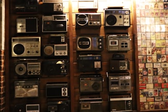 A collection of vintage radios displayed on a wooden wall, each with distinct designs and dials. To the right, an assortment of old album covers is arranged in a grid pattern.