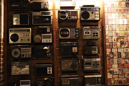 A collection of vintage radios displayed on a wooden wall, each with distinct designs and dials. To the right, an assortment of old album covers is arranged in a grid pattern.