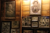 A collection of framed black and white photographs mounted on a wooden wall. The photos feature portraits and group images from various eras, showcasing people in formal and casual attire. The central focus includes two larger individual portraits above several smaller images.