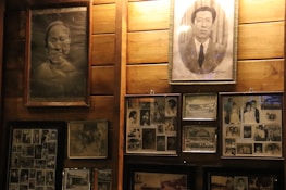A collection of framed black and white photographs mounted on a wooden wall. The photos feature portraits and group images from various eras, showcasing people in formal and casual attire. The central focus includes two larger individual portraits above several smaller images.