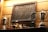 A wooden wall features a framed collection of vintage keys. Below the frame, several decorative objects including two silver trophies, and a series of carved wooden figurines and birds are displayed on a shelf. A modern trophy with a star design is also present.