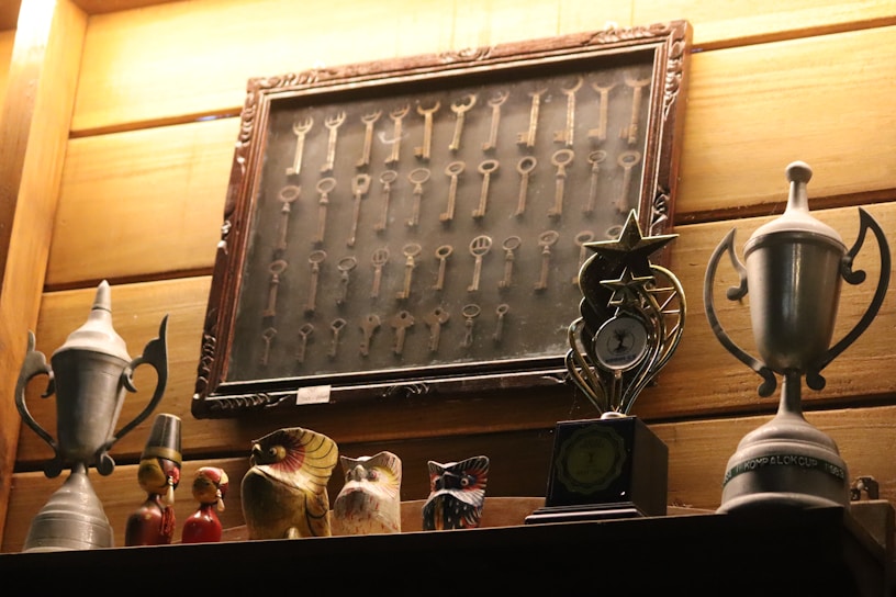 A wooden wall features a framed collection of vintage keys. Below the frame, several decorative objects including two silver trophies, and a series of carved wooden figurines and birds are displayed on a shelf. A modern trophy with a star design is also present.