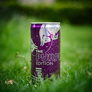 A purple can of energy drink is placed upright on a patch of green grass. The can is adorned with the logo of two charging bulls and marked with the words 'The Purple Edition'. Water droplets are visible on the surface, indicating it may be cold or recently exposed to moisture.