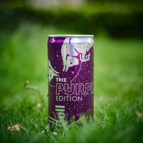 A purple can of energy drink is placed upright on a patch of green grass. The can is adorned with the logo of two charging bulls and marked with the words 'The Purple Edition'. Water droplets are visible on the surface, indicating it may be cold or recently exposed to moisture.