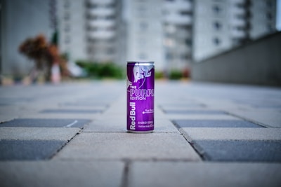 A can of Red Bull's Purple Edition energy drink is placed on a paved walkway, with a blurred urban background comprising buildings and greenery.