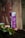 A can of Red Bull Purple Edition with acai berry flavor is placed on a textured surface surrounded by green leaves. The can is prominently purple with a reflective finish and some visible water droplets.