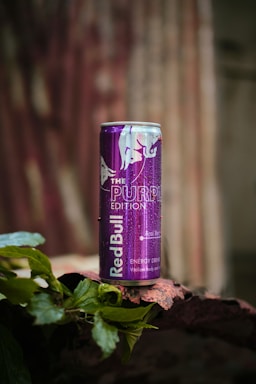 A can of Red Bull Purple Edition with acai berry flavor is placed on a textured surface surrounded by green leaves. The can is prominently purple with a reflective finish and some visible water droplets.
