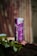 A can of Red Bull Purple Edition with acai berry flavor is placed on a textured surface surrounded by green leaves. The can is prominently purple with a reflective finish and some visible water droplets.