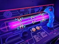 A close-up view of a retro-style arcade game interface, featuring a neon-lit control panel with vibrant purple and blue lights. A translucent blue joystick with a red button is positioned on the right side, surrounded by various geometric designs and arrows on the surface. The control panel includes instructions and two round yellow buttons.
