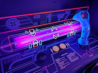 A close-up view of a retro-style arcade game interface, featuring a neon-lit control panel with vibrant purple and blue lights. A translucent blue joystick with a red button is positioned on the right side, surrounded by various geometric designs and arrows on the surface. The control panel includes instructions and two round yellow buttons.