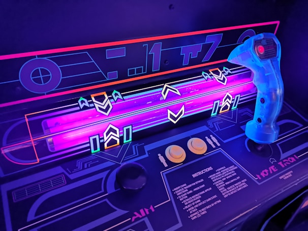 A close-up view of a retro-style arcade game interface, featuring a neon-lit control panel with vibrant purple and blue lights. A translucent blue joystick with a red button is positioned on the right side, surrounded by various geometric designs and arrows on the surface. The control panel includes instructions and two round yellow buttons.