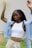 A person with braided hair is seen wearing a white crop top and beige high-waisted pants. Over the crop top, they have a light blue open shirt. Their arms are raised, and they appear to be outdoors with a blurred green background.