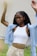 A person with braided hair is seen wearing a white crop top and beige high-waisted pants. Over the crop top, they have a light blue open shirt. Their arms are raised, and they appear to be outdoors with a blurred green background.