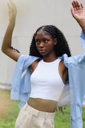 A person with braided hair is seen wearing a white crop top and beige high-waisted pants. Over the crop top, they have a light blue open shirt. Their arms are raised, and they appear to be outdoors with a blurred green background.