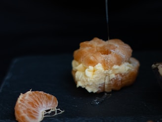 A cream puff filled with vanilla cream is being drizzled with syrup. Nearby, a slice of an orange rests on a dark surface, highlighting the contrast between the creamy dessert and the fresh fruit.