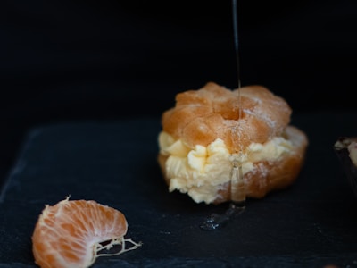 A cream puff filled with vanilla cream is being drizzled with syrup. Nearby, a slice of an orange rests on a dark surface, highlighting the contrast between the creamy dessert and the fresh fruit.