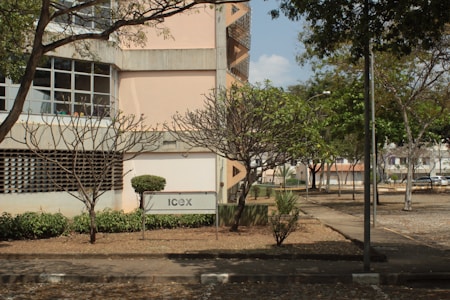 A building with concrete architecture is surrounded by a garden with leafless trees. A sign with 'Icex' is placed near a pathway. The area appears to be a part of a campus or institutional setting with paved walkways and shrubs.