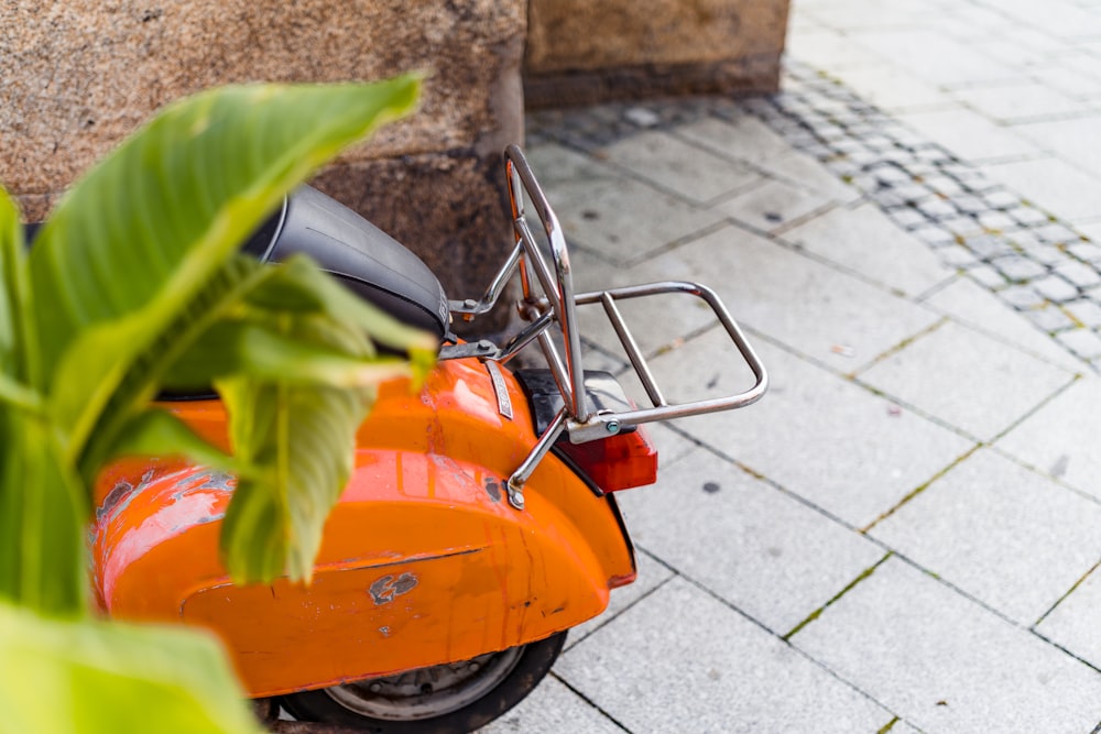 an orange scooter parked on a sidewalk next to a plant