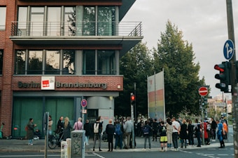 A group of people is gathered outside a brick building with large windows. The building is labeled 'Berlin Brandenburg'. The scene takes place on a city street, with trees and various street signs visible. There are several bicycles and scooters parked nearby. The group appears to be engaged in some form of public gathering or event.