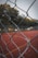 A wire mesh fence is in focus in the foreground, with a sports court featuring red surface and white boundary lines in the background. The scene is surrounded by trees, suggesting it is located outdoors in a park.