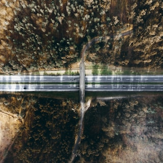An aerial view of a highway cutting through a dense forest area, with intersecting roads and visible greenery. The road is flanked by trees, displaying a serene and remote atmosphere.