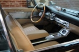 A classic car interior with beige leather seats and a wooden steering wheel. The dashboard features vintage controls and a retro design. Brick walls are visible through the windows, suggesting an indoor setting.