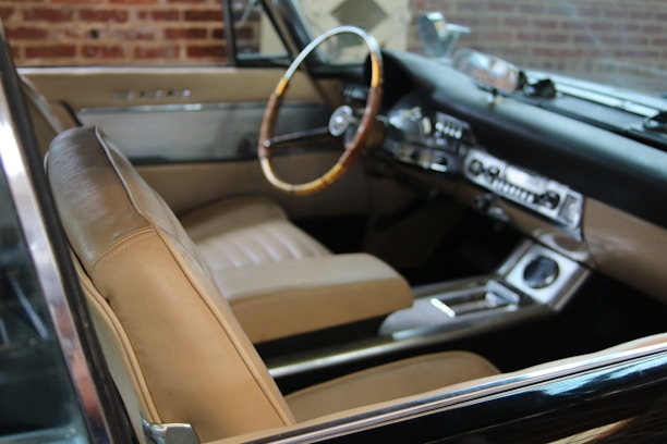 A classic car interior with beige leather seats and a wooden steering wheel. The dashboard features vintage controls and a retro design. Brick walls are visible through the windows, suggesting an indoor setting.