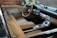 A classic car interior with beige leather seats and a wooden steering wheel. The dashboard features vintage controls and a retro design. Brick walls are visible through the windows, suggesting an indoor setting.