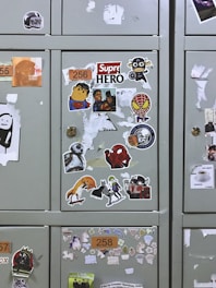 Several lockers covered with various stickers, including cartoon characters like superheroes, a robot, and other pop culture references. The lockers have patches where stickers have been removed, and each features a number label.