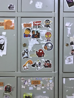 Several lockers covered with various stickers, including cartoon characters like superheroes, a robot, and other pop culture references. The lockers have patches where stickers have been removed, and each features a number label.