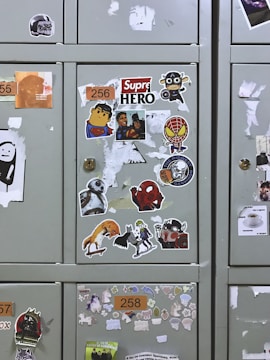 Several lockers covered with various stickers, including cartoon characters like superheroes, a robot, and other pop culture references. The lockers have patches where stickers have been removed, and each features a number label.