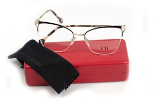 A pair of eyeglasses with a sleek design lies on a red glasses case. The case is embossed with the initials CH and the name Carolina Herrera. A black cleaning cloth is partially covering the case.