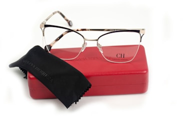 A pair of eyeglasses with a sleek design lies on a red glasses case. The case is embossed with the initials CH and the name Carolina Herrera. A black cleaning cloth is partially covering the case.