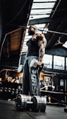A muscular individual stands in a dimly lit gym holding large dumbbells. The gym has industrial elements with a high ceiling and exposed beams. Various gym equipment and people can be seen in the background.