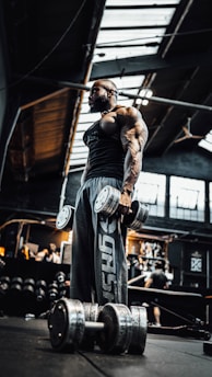 A muscular individual stands in a dimly lit gym holding large dumbbells. The gym has industrial elements with a high ceiling and exposed beams. Various gym equipment and people can be seen in the background.