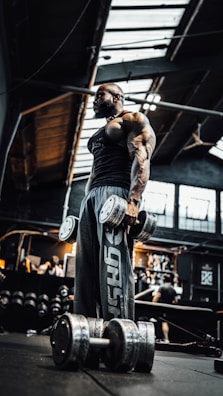 A muscular individual stands in a dimly lit gym holding large dumbbells. The gym has industrial elements with a high ceiling and exposed beams. Various gym equipment and people can be seen in the background.