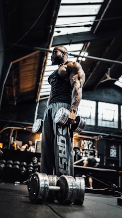 A muscular individual stands in a dimly lit gym holding large dumbbells. The gym has industrial elements with a high ceiling and exposed beams. Various gym equipment and people can be seen in the background.