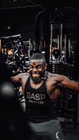 A muscular man is intensely working out in a gym, wearing a black gym tank top with the word 'GASP' on it. He is performing a cable exercise, with a strong expression of effort on his face. The gym is filled with various fitness equipment in the background.