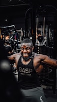 A muscular man is intensely working out in a gym, wearing a black gym tank top with the word 'GASP' on it. He is performing a cable exercise, with a strong expression of effort on his face. The gym is filled with various fitness equipment in the background.