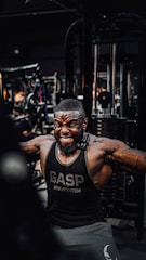 A muscular man is intensely working out in a gym, wearing a black gym tank top with the word 'GASP' on it. He is performing a cable exercise, with a strong expression of effort on his face. The gym is filled with various fitness equipment in the background.