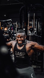 A muscular man is intensely working out in a gym, wearing a black gym tank top with the word 'GASP' on it. He is performing a cable exercise, with a strong expression of effort on his face. The gym is filled with various fitness equipment in the background.