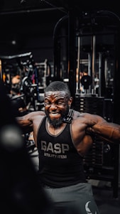 A muscular man is intensely working out in a gym, wearing a black gym tank top with the word 'GASP' on it. He is performing a cable exercise, with a strong expression of effort on his face. The gym is filled with various fitness equipment in the background.