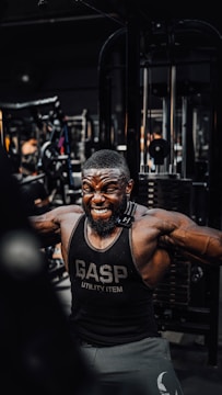 A muscular man is intensely working out in a gym, wearing a black gym tank top with the word 'GASP' on it. He is performing a cable exercise, with a strong expression of effort on his face. The gym is filled with various fitness equipment in the background.