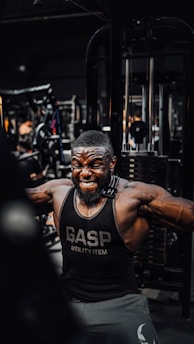 A muscular man is intensely working out in a gym, wearing a black gym tank top with the word 'GASP' on it. He is performing a cable exercise, with a strong expression of effort on his face. The gym is filled with various fitness equipment in the background.