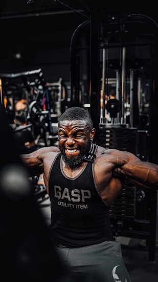 A muscular man is intensely working out in a gym, wearing a black gym tank top with the word 'GASP' on it. He is performing a cable exercise, with a strong expression of effort on his face. The gym is filled with various fitness equipment in the background.
