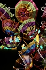 Colorful wire sculptures shaped like geometric baskets hang against a dark background. The intricate patterns of the baskets are created by using vibrant metal wires twisted and connected to form detailed structures.