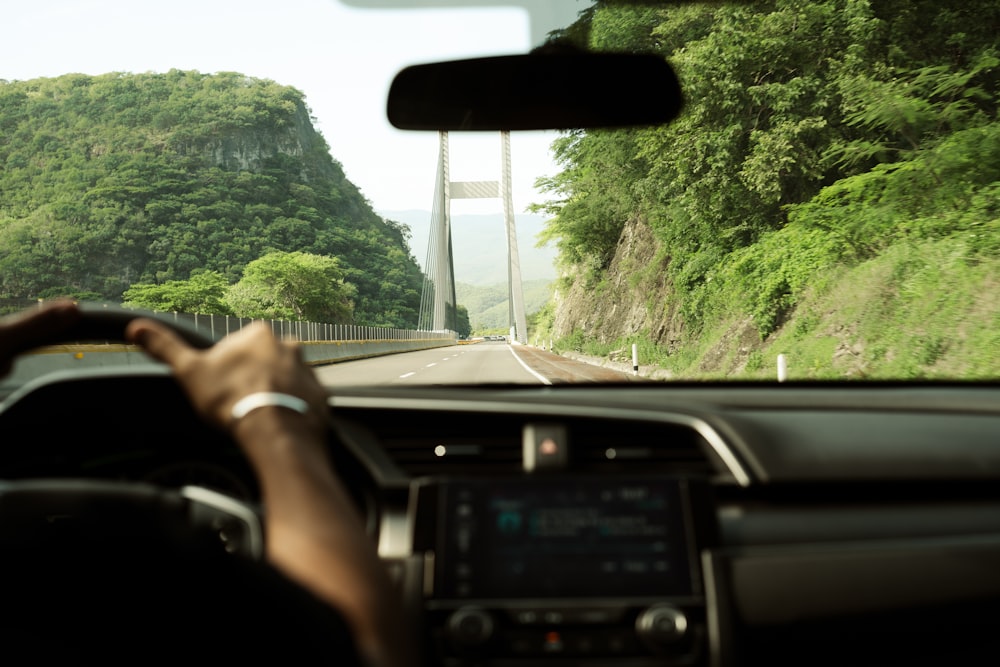 a man driving a car down a road next to a lush green hillside