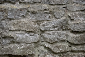 A textured stone wall constructed with irregularly shaped, rough stones stacked on top of each other. The stones vary in size and are interspersed with thin lines of mortar.