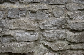 A textured stone wall constructed with irregularly shaped, rough stones stacked on top of each other. The stones vary in size and are interspersed with thin lines of mortar.